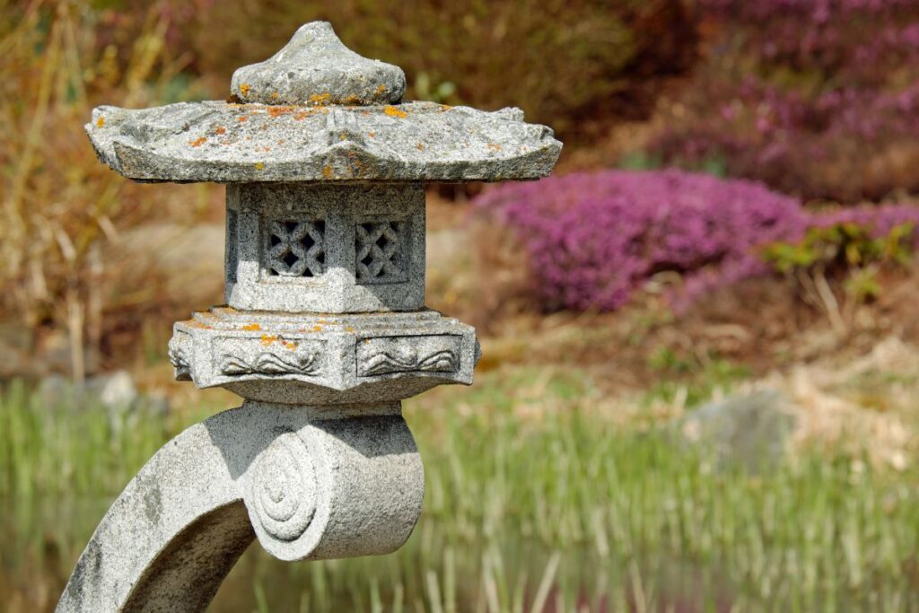Granite lantern post with intricate details, surrounded by vibrant pink flowering shrubs and greenery, enhancing traditional New England landscape aesthetics.
