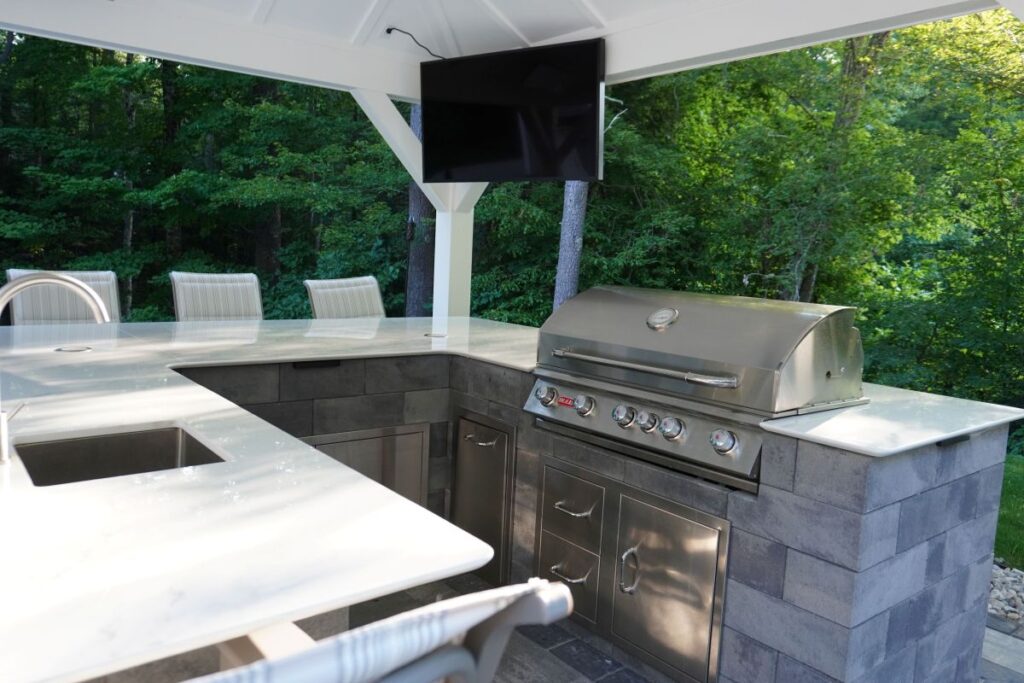 Granite outdoor kitchen with stainless steel grill, marble countertop, and seating, surrounded by lush greenery, showcasing modern outdoor living features.