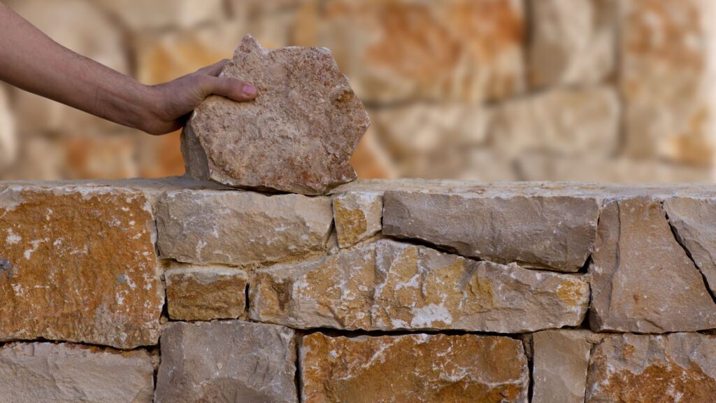 Hand placing stone onto a partially constructed retaining wall, showcasing hardscaping techniques for durable landscape features.