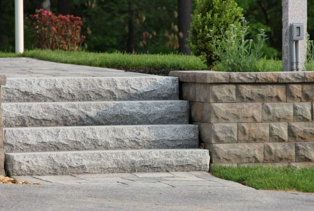 Granite steps leading up to a patio, surrounded by a stone retaining wall and landscaped greenery, emphasizing durable hardscaping features for outdoor spaces.