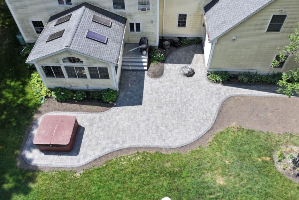 Patio with paver stones, hot tub, and landscaped yard adjacent to a home, showcasing summer outdoor living features.