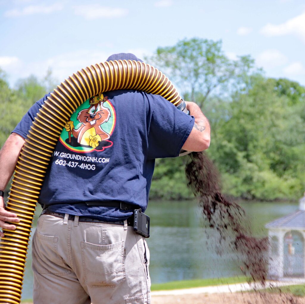 Landscaper using a mulch blower to distribute dark hardwood mulch, promoting healthy landscapes and soil enrichment, with a river and greenery in the background.