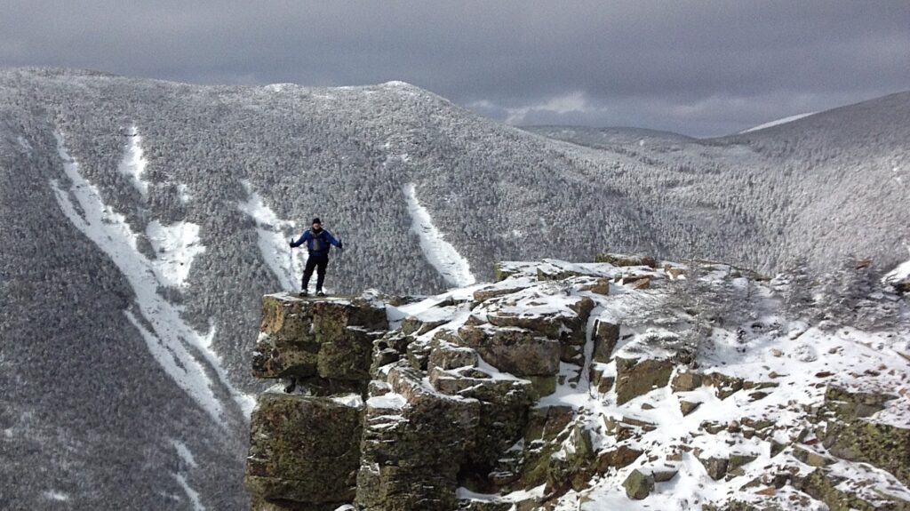 Man standing on rocky outcrop in snowy mountain landscape, showcasing New Hampshire's natural granite features and rugged terrain.