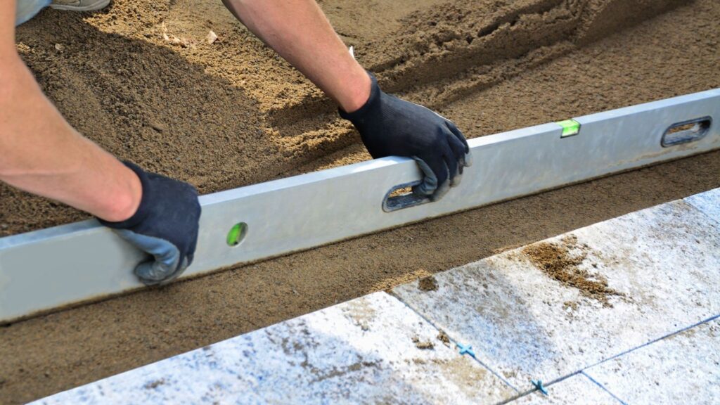 Person leveling sand for hardscape installation, focusing on patio construction, emphasizing summer landscaping projects.