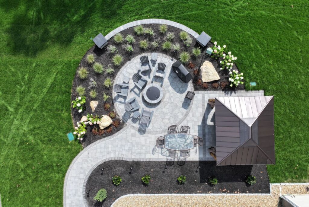 Aerial view of a landscaped patio with a circular fire pit, surrounded by seating, decorative stones, and garden beds, showcasing hardscape design principles for outdoor living spaces.