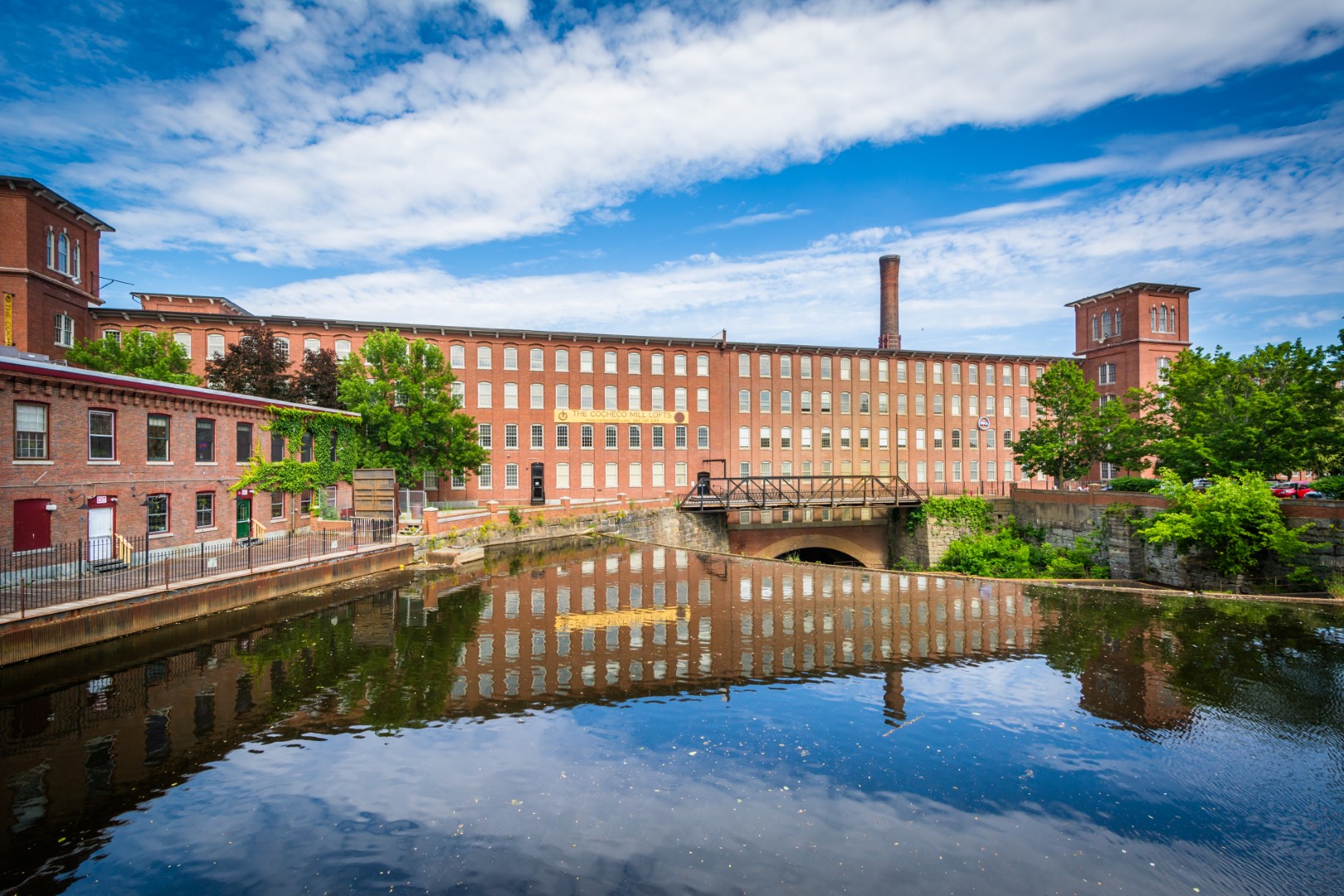 Historic brick mill building reflecting in water, surrounded by greenery and blue sky, representing Manchester, NH's landscape and architectural heritage.
