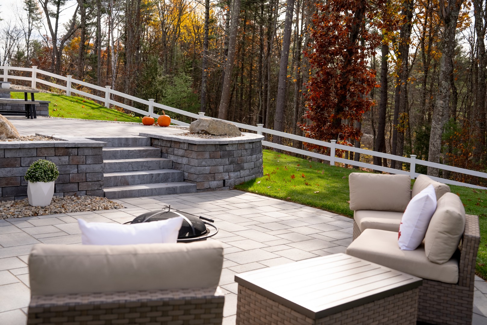 Outdoor living space featuring a stone patio with seating area, fire pit, steps leading to a raised section, and decorative pumpkins, surrounded by trees and a white fence, illustrating durable materials and design elements from Groundhog Landscaping in Milford, NH.