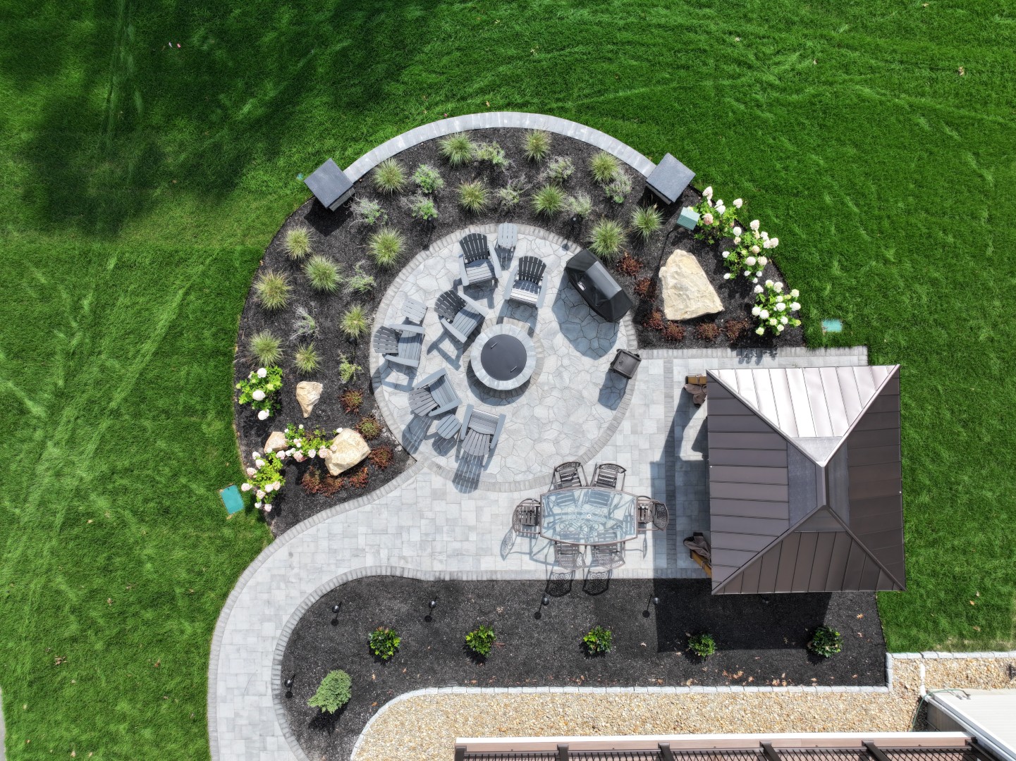 Custom outdoor living space featuring a circular stone patio with seating around a fire pit, surrounded by landscaped greenery and a gazebo, designed for New Hampshire homes by Groundhog Landscaping.