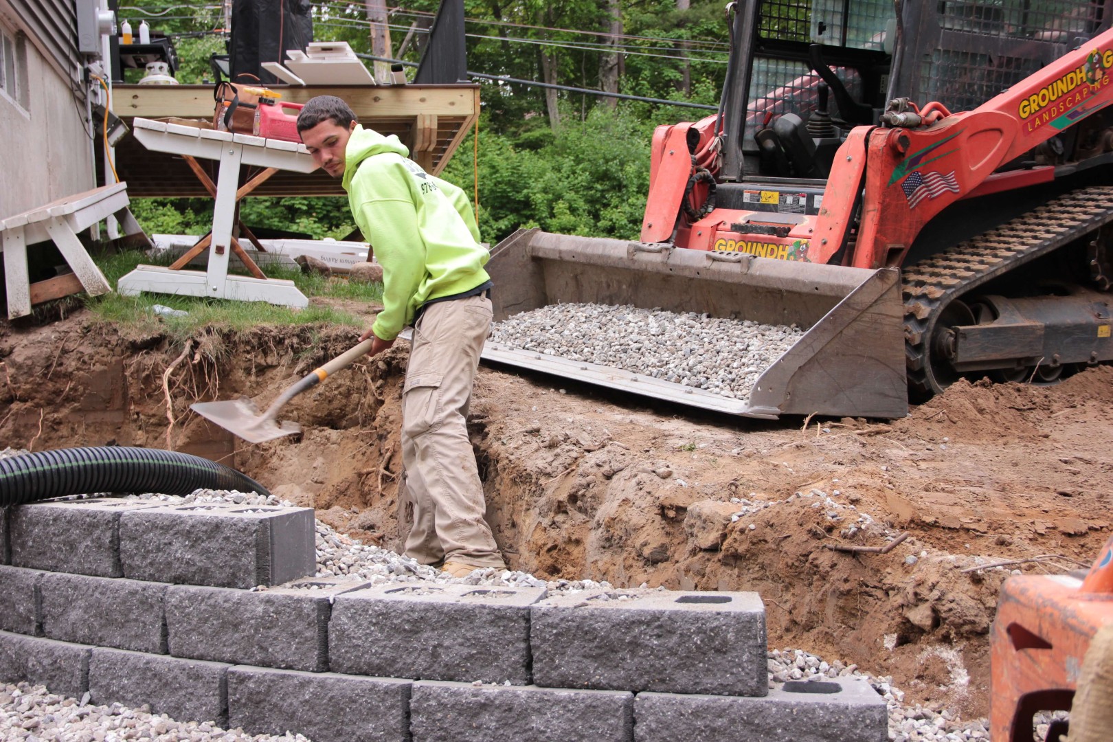 Worker using a shovel to move gravel near a wall of gray pavers, with a Groundhog Landscaping machine in the background, illustrating the construction process for landscaping projects.