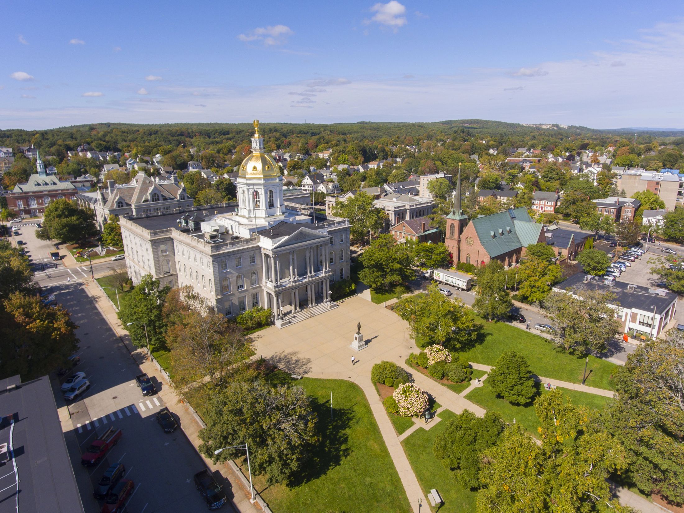Aerial view of Concord, NH, showcasing the New Hampshire State House with its gold dome, surrounded by vibrant greenery, historic buildings, and community spaces, emphasizing the city's blend of natural beauty and cultural significance.