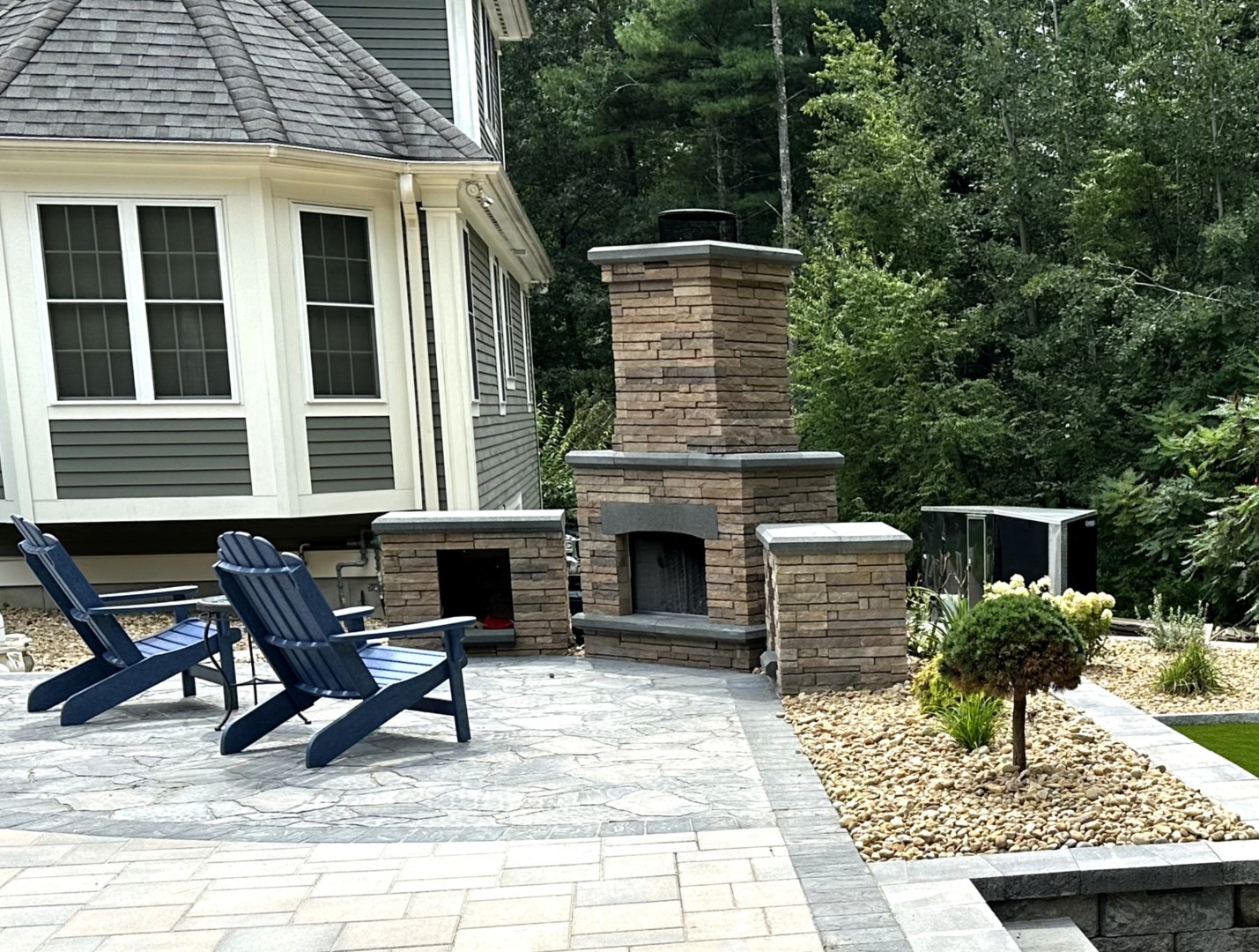 Outdoor living space featuring stone fire pit, blue Adirondack chairs, and landscaped garden with greenery, designed for comfort and entertainment by Groundhog Landscaping.