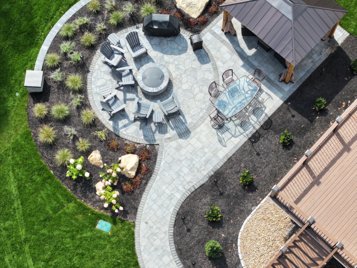 Aerial view of a beautifully designed outdoor living space featuring a stone patio, circular fire pit surrounded by Adirondack chairs, a dining table, and a gazebo, with landscaped gardens and decorative rocks, showcasing Groundhog Landscaping's expertise in landscape construction in Salem, NH.