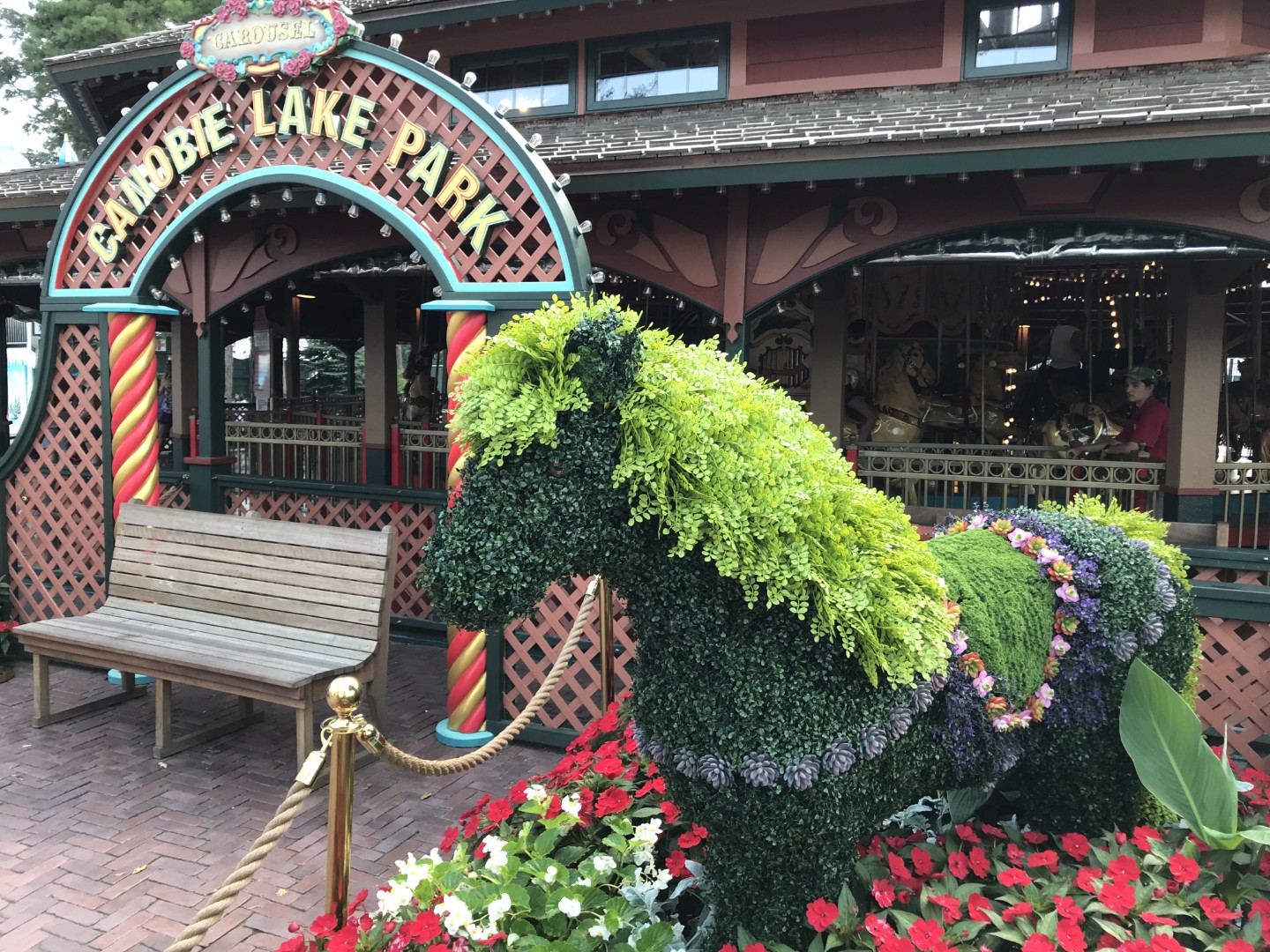 Topiary horse sculpture adorned with vibrant flowers at Canobie Lake Park entrance, surrounded by colorful flower beds and a wooden bench.