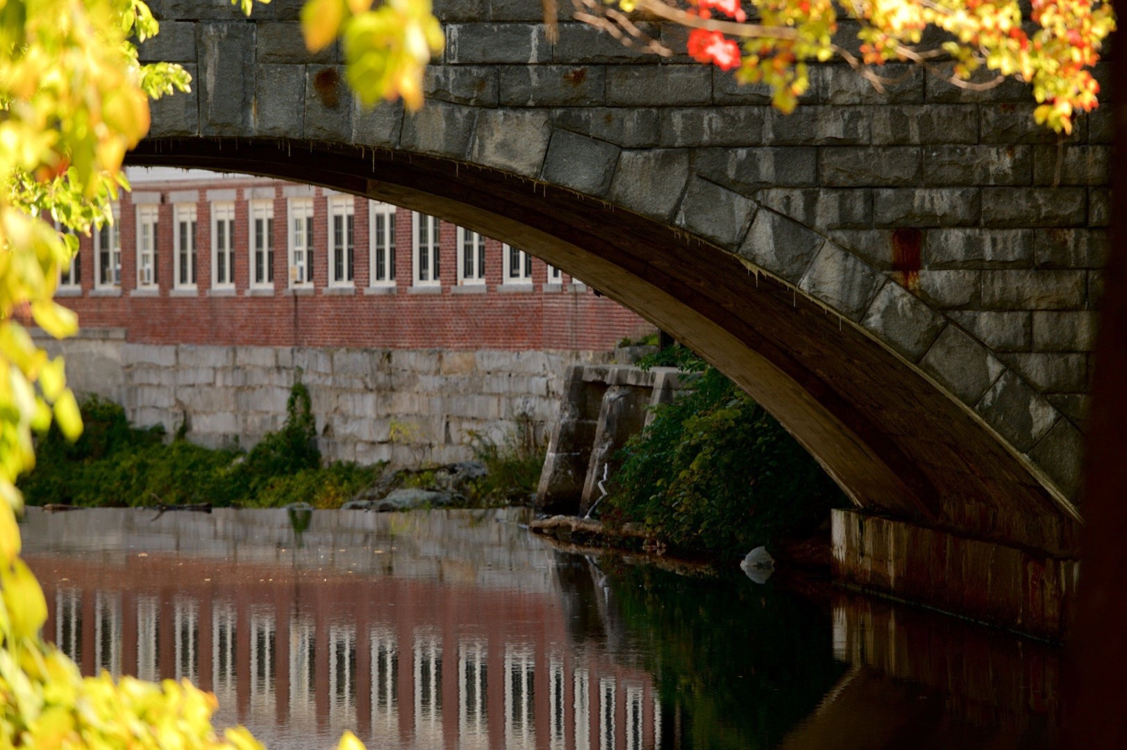 Stone arch bridge reflecting in water, surrounded by greenery, with historical brick building in the background, representing Milford, NH landscape.