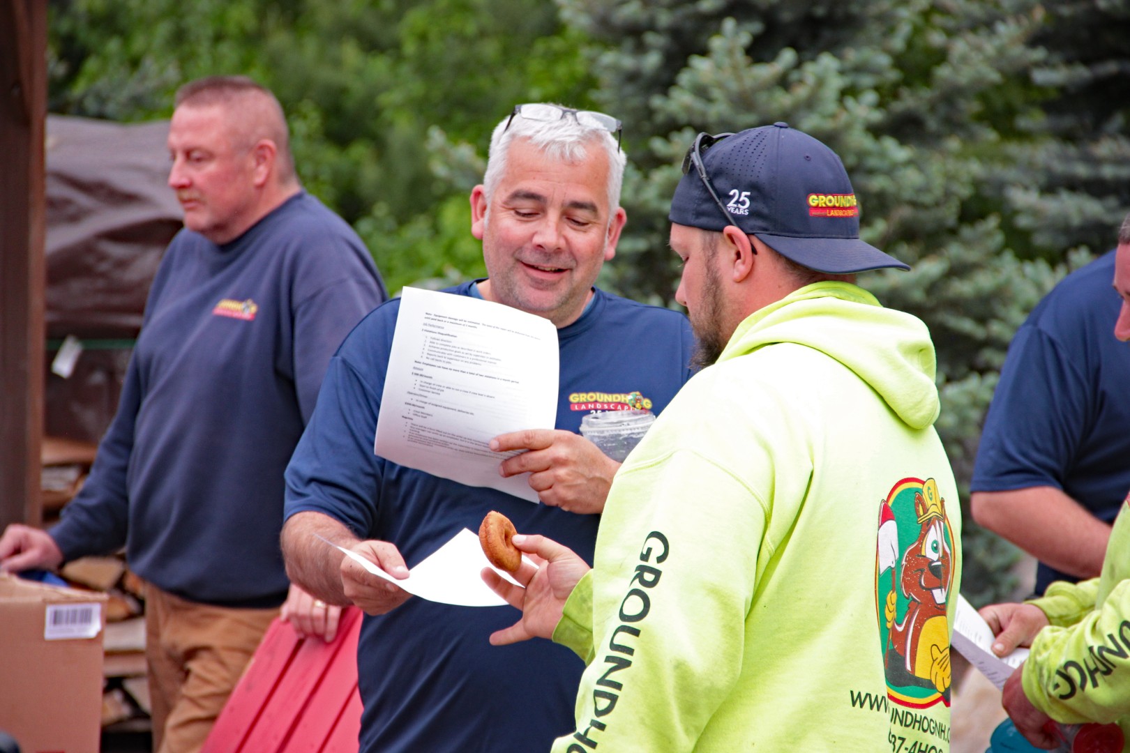 Groundhog Landscaping team members discussing project details, with one holding a document and another holding a donut, showcasing collaboration and team engagement in Milford, NH.