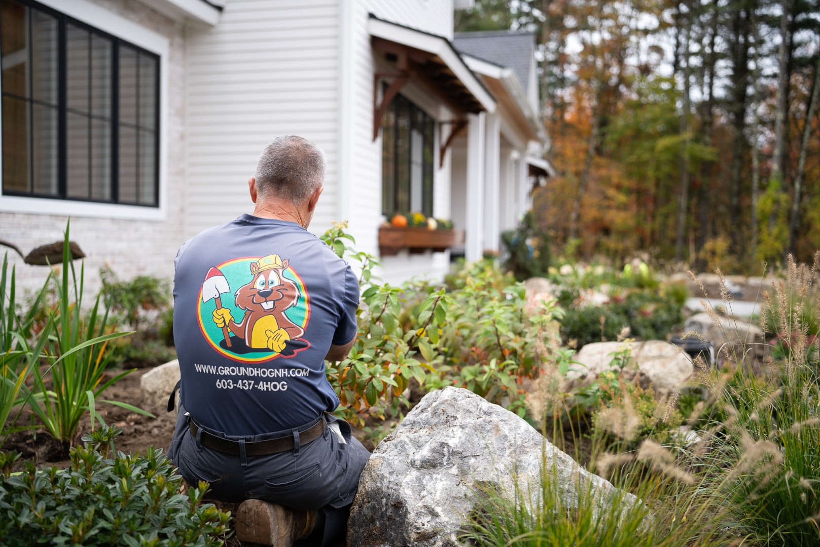 Groundhog Landscaping professional tending to vibrant garden plants, wearing branded shirt with cartoon groundhog logo, surrounded by rocks and landscaping elements.