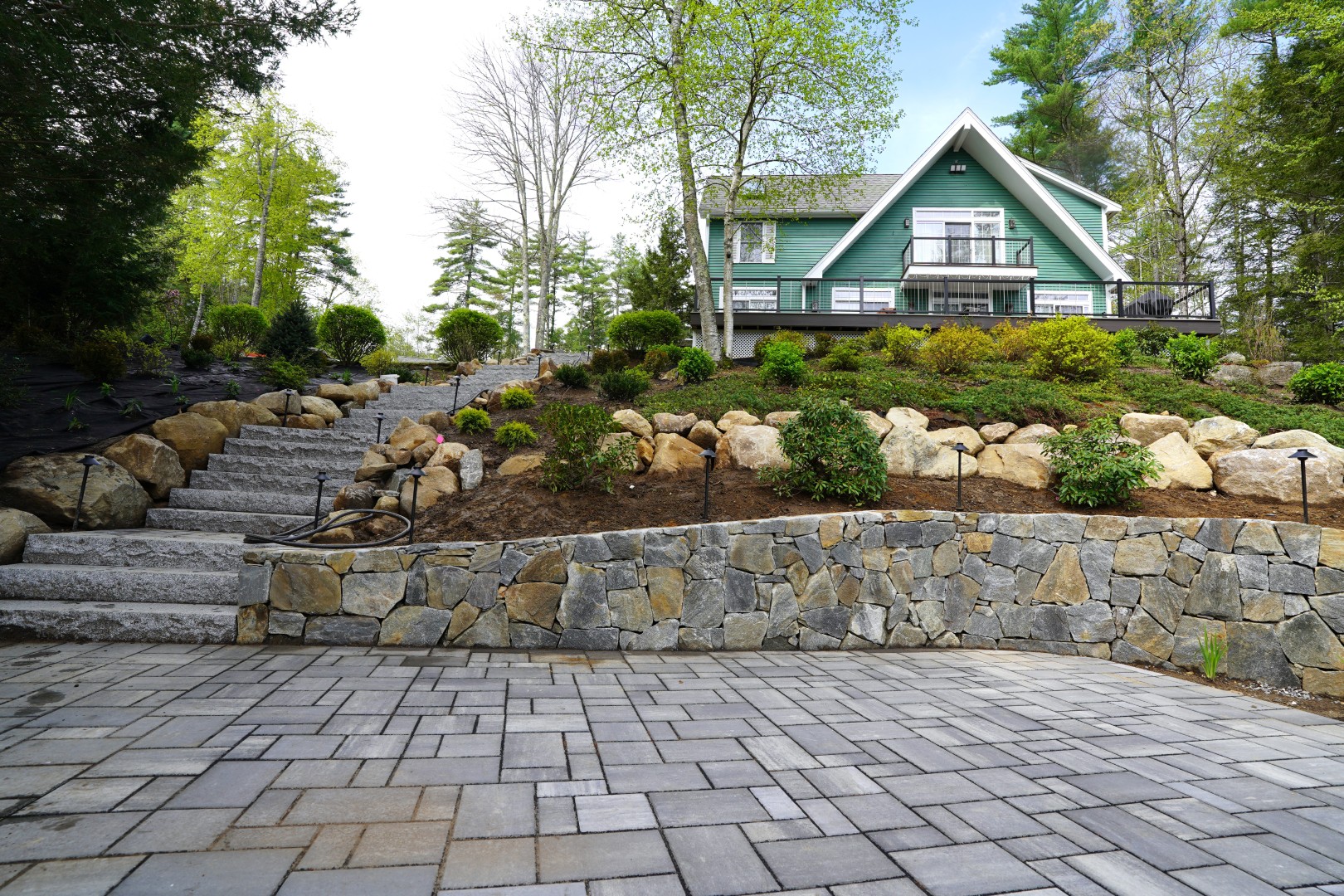 Stone retaining wall and steps leading to a landscaped garden with greenery and a modern house in Exeter, NH, showcasing effective erosion management and aesthetic enhancement.