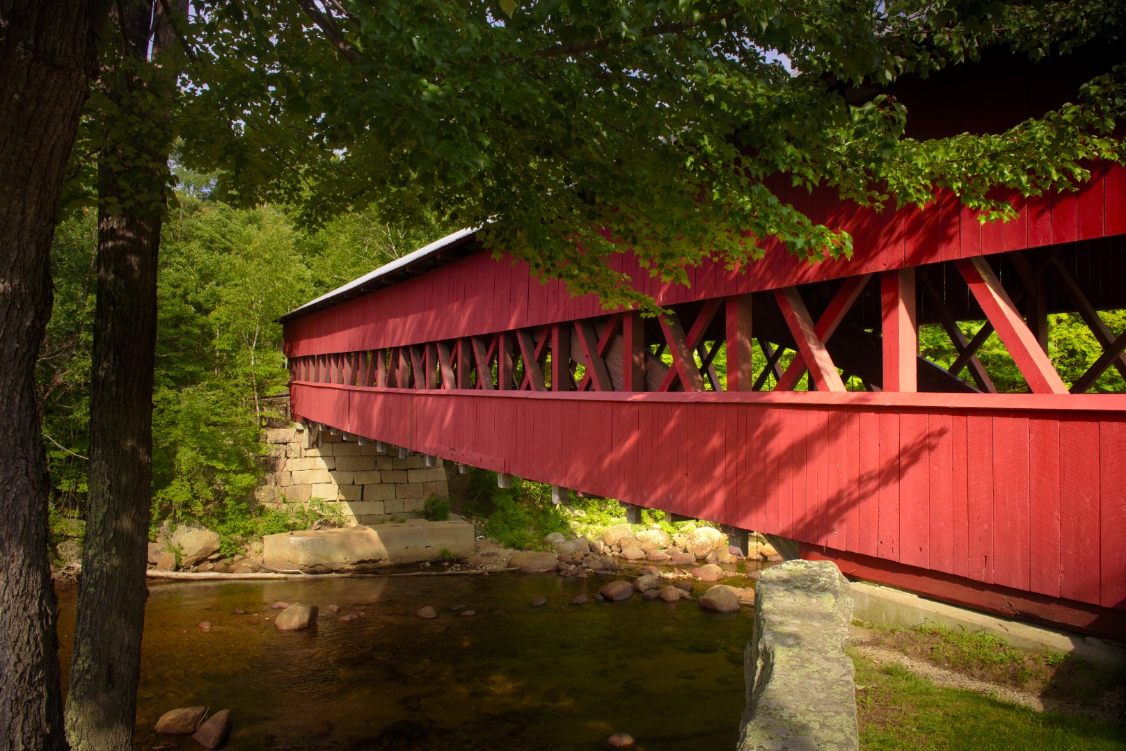 Red covered bridge surrounded by lush greenery, spanning a clear stream in Chester, NH, showcasing local historical architecture and natural beauty.