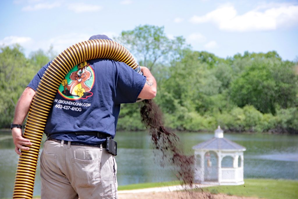 Man using bark blowing equipment to apply mulch near a lake, showcasing Groundhog Landscaping's efficient landscaping services.