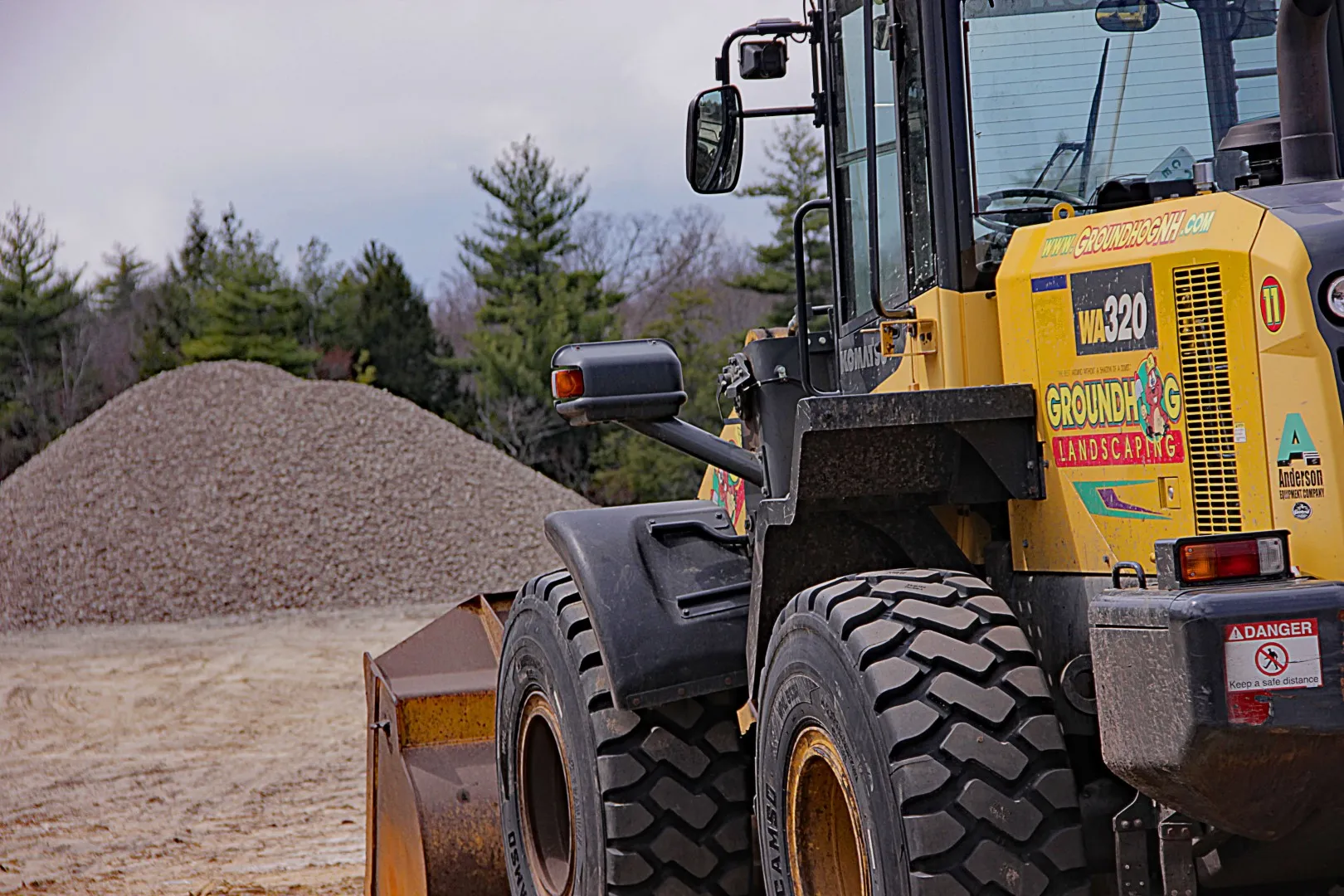 Komatsu WA320 loader with Groundhog Landscaping branding, positioned near a large pile of gravel in a landscape materials delivery setting in Salem, NH.