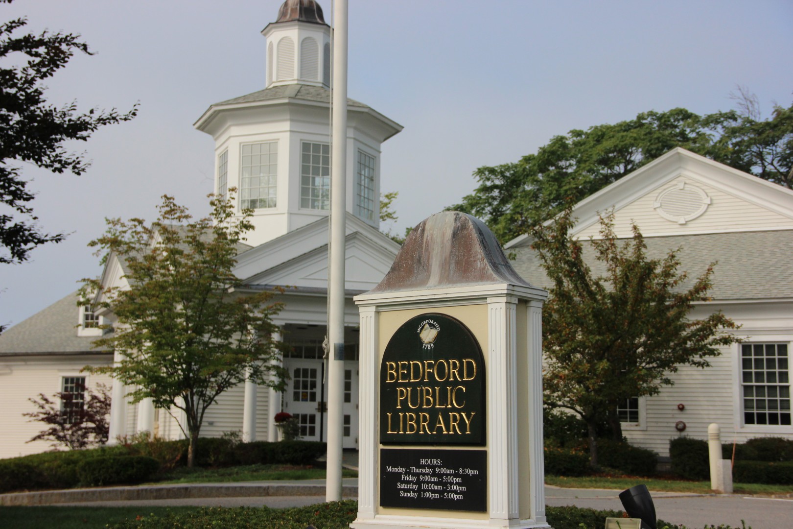 Bedford Public Library exterior with sign displaying hours and services, surrounded by trees and landscaping in Bedford, NH.