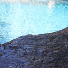 Close-up of a textured stone edge beside a sparkling blue swimming pool, highlighting the hardscaping features of the Londonderry Pool Patio project by Groundhog Landscaping.